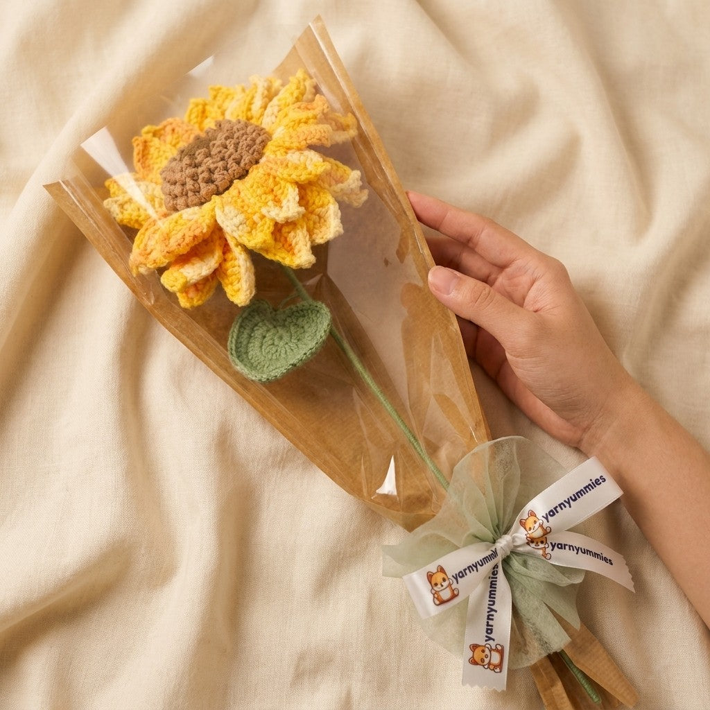 Hand holding a transparent flower sleve with a knitted sunflower and green leaf on a beige fabric background.