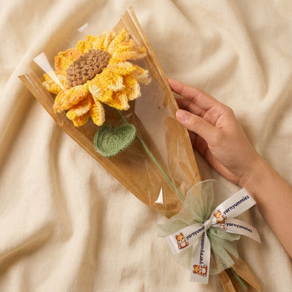 Hand holding a transparent flower sleve with a knitted sunflower and green leaf on a beige fabric background.
