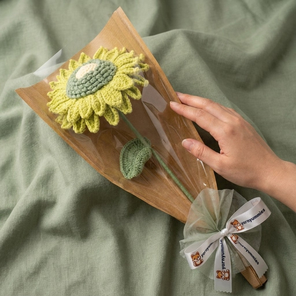 Hand holding a knitted sunflower toy wrapped in brown paper with a white ribbon on a green fabric background.