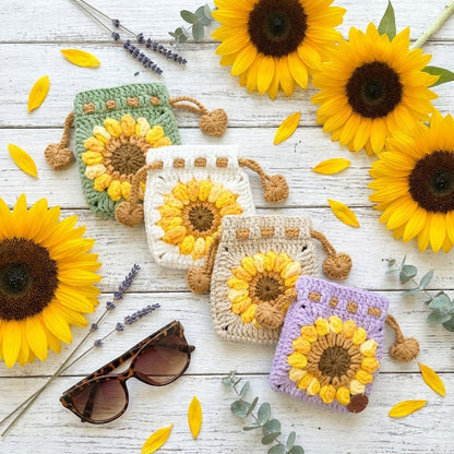 4 crocheted sunflower designed pouches on a wooden surface with sunflowers and sunglasses.