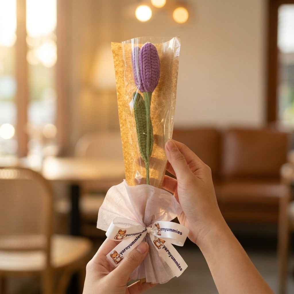 Hand holding a small bouquet of purple crocheted flower wrapped in clear plastic with a white ribbon against a blurred indoor background.