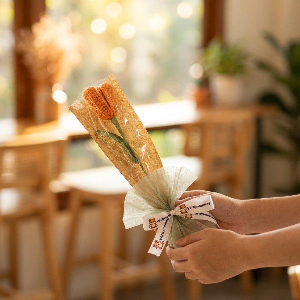 Bouquet of crocheted tulip wrapped in brown paper with a white ribbon held by a person in a blurred indoor setting.