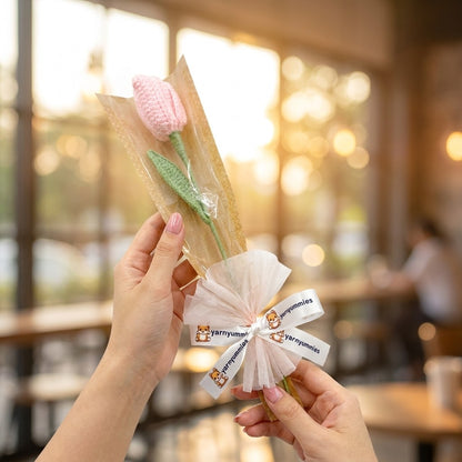 Two hands holding a bouquet of crocheted pink tulip with 'Yarnyummies' branding against a blurred indoor background.