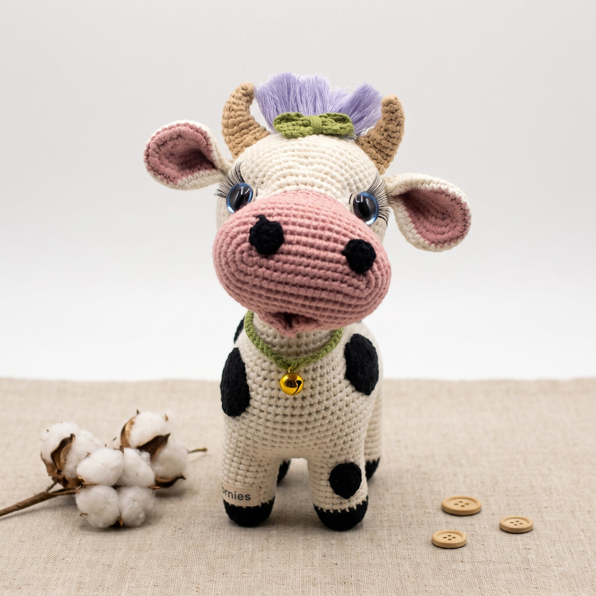 A handmade crocheted plush black and white cow toy by YarnYummies standing on a beige fabric surface with cotton bolls and wooden buttons nearby. The toy has a cream body, pink snout and ears, black spots, tan horns, a purple horn tuft, and a green collar with a small gold bell.