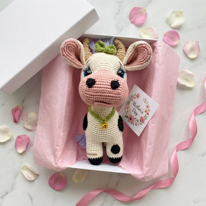 Crocheted cow plushie in a box with pink ribbon and Mother's Day card on a marble surface.